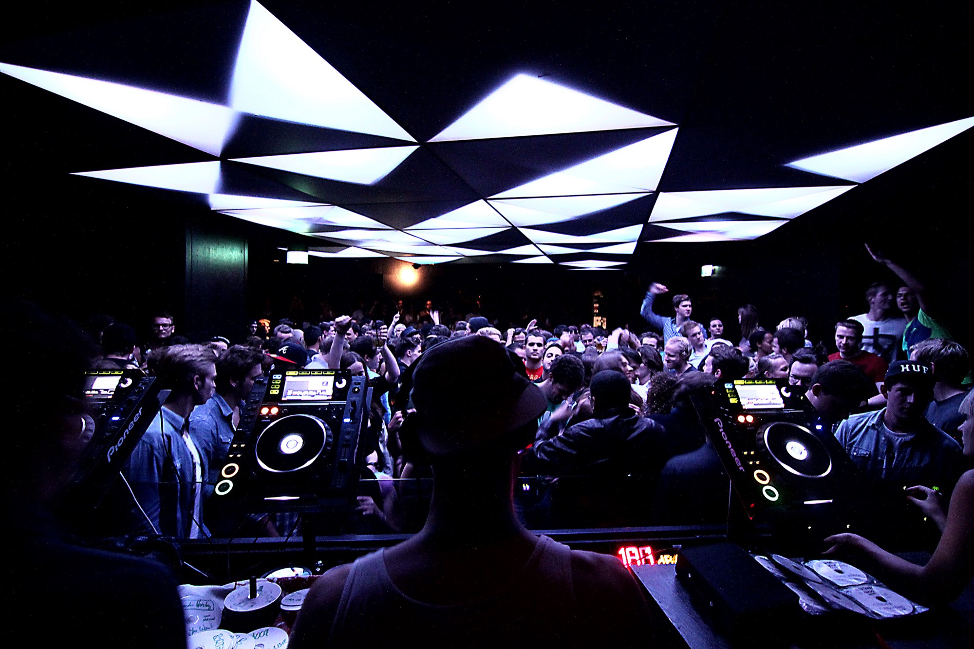 A DJ booth facing a packed dance floor under white light panels